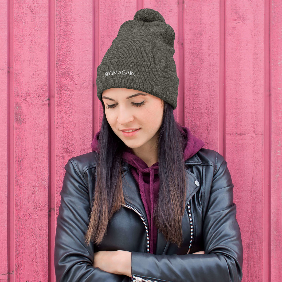 Woman standing in front of pink wall wearing a leather jacket and Begin Again beanie hat.